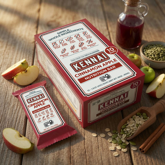 A box and individually wrapped Kenai Cinnamon Apple Nutrition Bar are displayed on a rustic wooden table with sliced apples, oats, pumpkin seeds, cinnamon sticks, and a small bottle of honey.